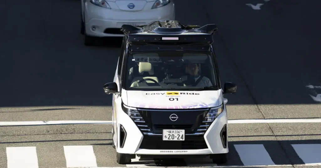 A front-facing, eye-level shot shows a white Nissan Easy Ride autonomous vehicle moving along a crosswalk. The vehicle has a black roof and a sensor array on top. The front of the vehicle features the Nissan logo, a black grille, and the words "Easy Ride by Nissan" in blue and yellow.