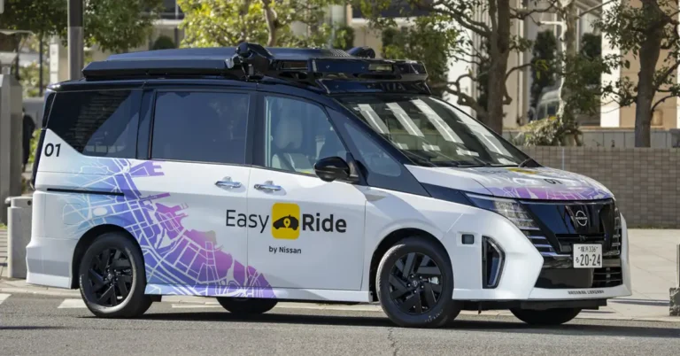 A white Nissan Serena minivan with black accents and black wheels, equipped with a roof rack and sensors, parked on a city street.