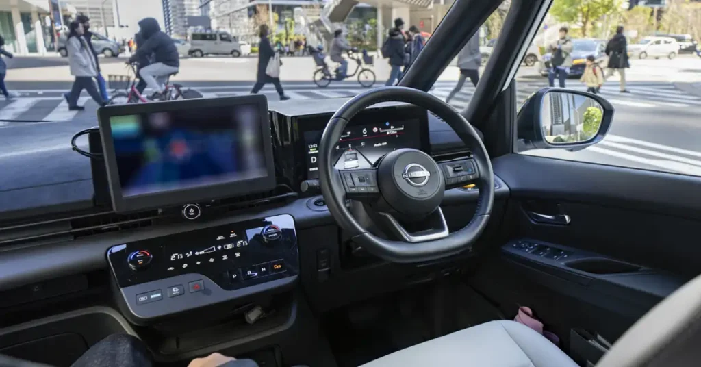 Interior view of a Nissan car, showing the dashboard, steering wheel, and digital displays, with a view through the windshield of a city street with pedestrians and cyclists crossing a zebra crossing. The dashboard is sleek and modern, featuring a large central display screen and a smaller screen mounted on top.