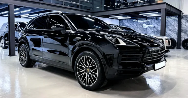 A sleek, black Porsche Cayenne SUV is parked inside a modern showroom. The car's glossy finish reflects the overhead lighting, accentuating its aerodynamic design.