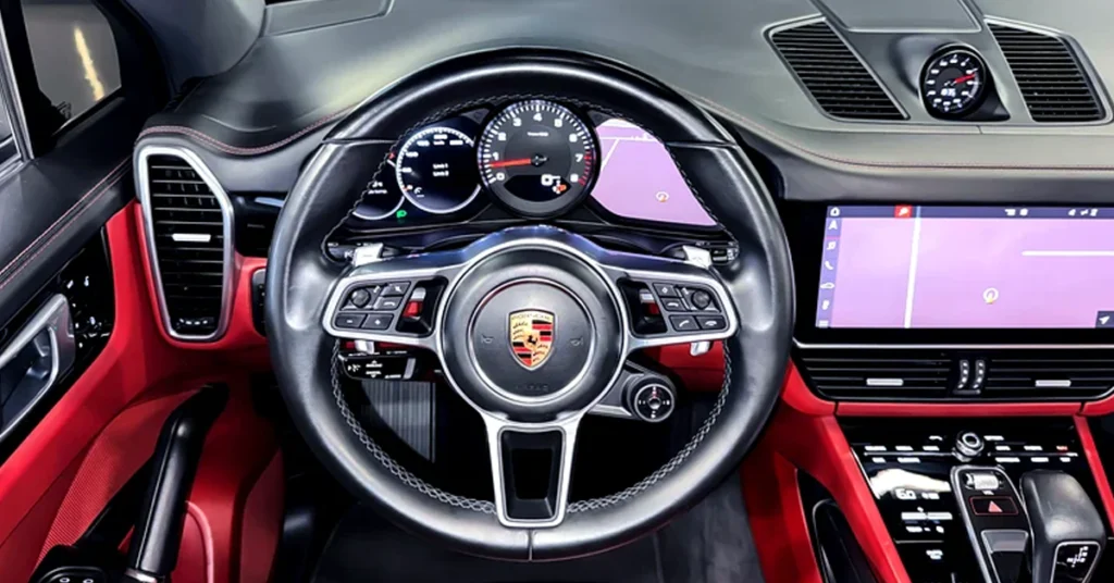 Close-up shot of the interior of a Porsche Cayenne focusing on the steering wheel and dashboard. The steering wheel is black leather with silver accents and the Porsche logo in the center. The dashboard is black with red leather accents and features a digital display screen, analog gauges, and various buttons and controls. The overall style is modern and luxurious.