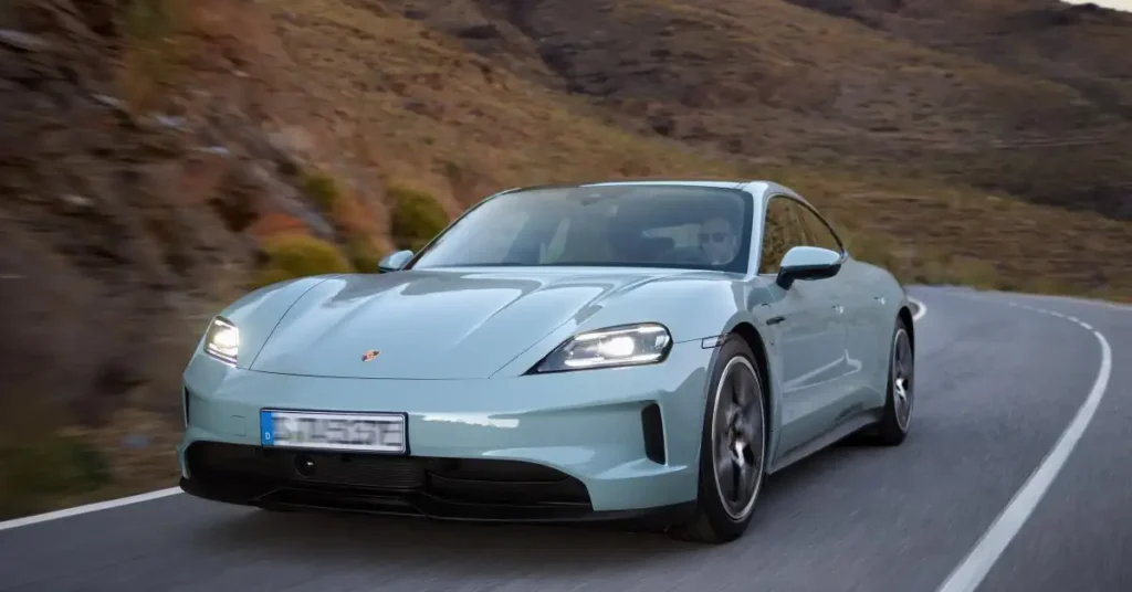 A light blue Porsche Taycan driving on a road with mountains in the background. The car is moving towards the viewer, and the road curves slightly to the right.