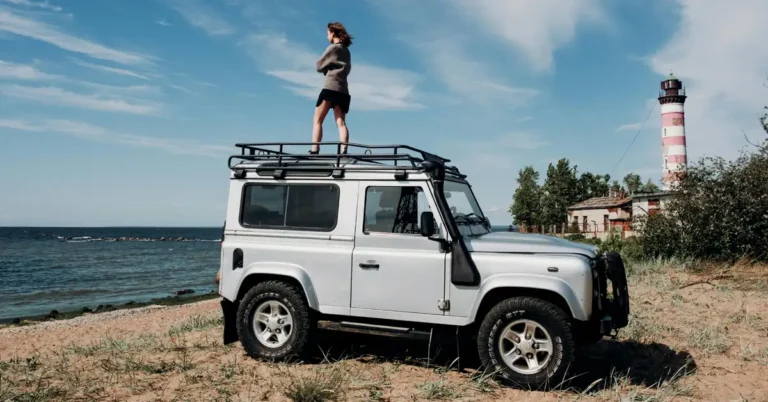 A full shot captures a Caucasian woman, around 25 years old, standing on the roof rack of a silver Land Rover Defender on a sandy beach.