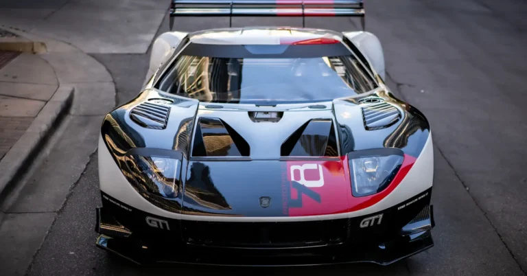 A front-facing, eye-level shot captures a Ford GT 1 race car parked on a city street. The car features a striking color scheme of white, red, and black, with the number "78" prominently displayed on its hood. The car's design includes a large rear wing, air vents on the hood, and a low-slung profile