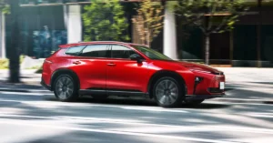 A red Toyota Crown Estate 2025 SUV is in motion on a city street, with blurred trees and buildings in the background. The vehicle has a sleek, modern design with a sloping roofline and dark-colored wheels.