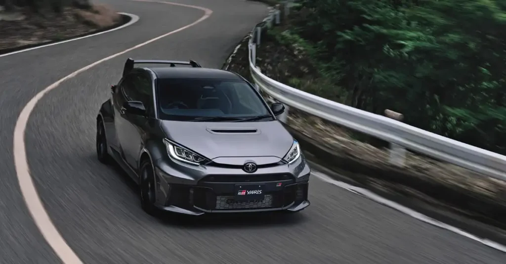 A gray Toyota GR Yaris races along a winding asphalt road, captured in a dynamic, slightly low-angle shot. The car, a compact hatchback, features a black roof and a prominent rear spoiler, enhancing its sporty appearance. Its headlights are illuminated, and the "GR Yaris" badge is visible on the front grille. The road curves gently, marked with a white line, suggesting a mountain pass or scenic route.