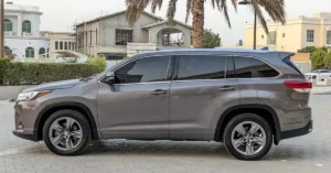 A gray Toyota Highlander SUV is parked on a paved surface, with a side view of the vehicle visible. The car has tinted windows and silver rims. In the background, there are several buildings, including one under construction with scaffolding.