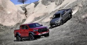 Two Toyota Hilux Hybrid 48V pickup trucks, one red and one gray, are positioned on a steep, rocky hillside. The red truck is in the foreground, angled slightly towards the viewer, showcasing its front design.