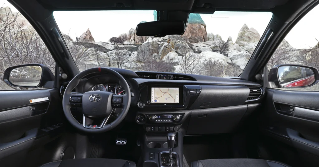 Interior view of a Toyota Hilux GR Sport, featuring a black dashboard, steering wheel with red accents, and a central infotainment screen displaying a navigation map. The gear stick is visible in the center console, and the exterior view through the windshield shows a landscape with rock formations and sparse vegetation.
