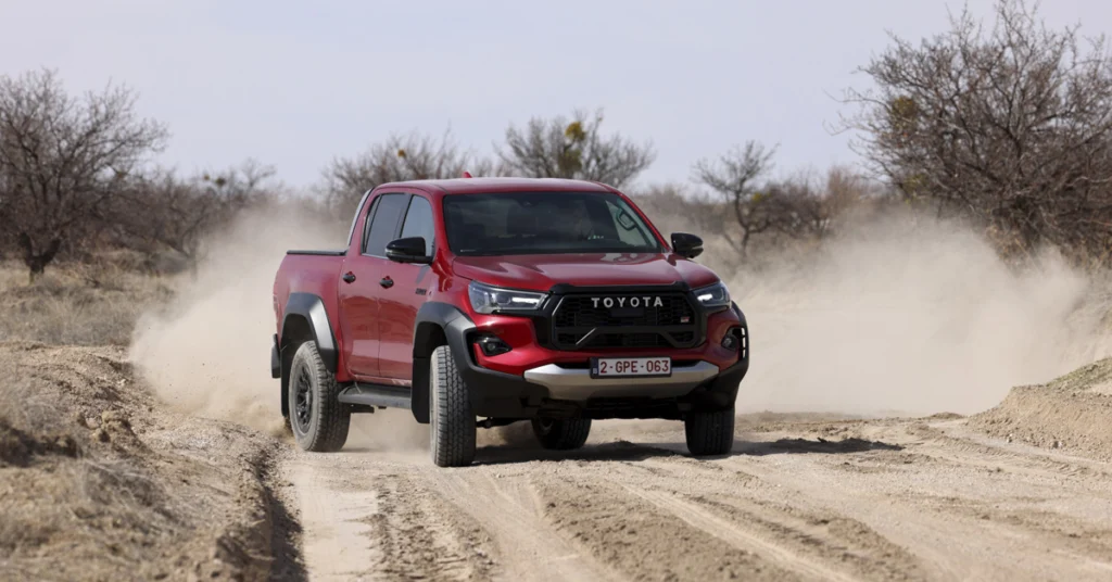A red Toyota Hilux GR Sport pickup truck is driving on a dirt road, kicking up dust. The truck is facing the camera and is slightly angled to the left. The background is a dry, arid landscape with sparse vegetation and bare trees.