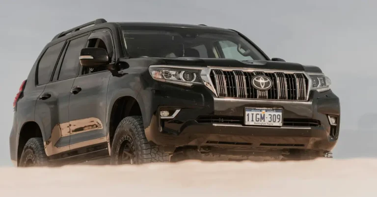 A black Toyota Land Cruiser Prado is shown in a medium shot, positioned at a low angle. The vehicle is partially submerged in sand, with the license plate visible, reading "1IGM-309". The car is surrounded by a sandy environment, and the sky is overcast. The lighting is soft and diffused, creating a muted color palette. The image is captured with a shallow depth of field, focusing on the front of the car.