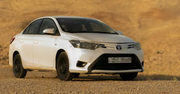 A white 2014 Toyota Yaris sedan is parked on a gravelly surface with a tan, hilly background. The car is positioned at a slight angle, facing towards the left of the frame.