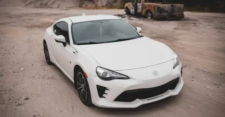 A medium shot captures a white Toyota 86 sports car parked on a dirt road, with a burnt-out truck in the background. The car is positioned at a slight angle, showcasing its sleek design and sporty features.