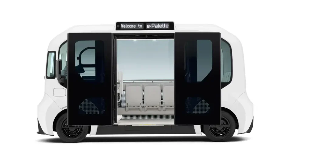 A digital rendering of a white e-Palette vehicle with its doors open, set against a stark white background. The vehicle has a boxy, futuristic design with rounded edges and a large, open doorway revealing the interior. Inside, there are several white seats arranged in a row.
