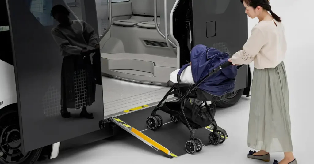 A medium shot captures a Japanese woman, around 30 years old, with fair skin and black hair tied back in a ponytail, dressed in a long green skirt and a beige long-sleeved shirt, pushing a black stroller with a blue cover up a ramp into a black and white Toyota e-Palette.