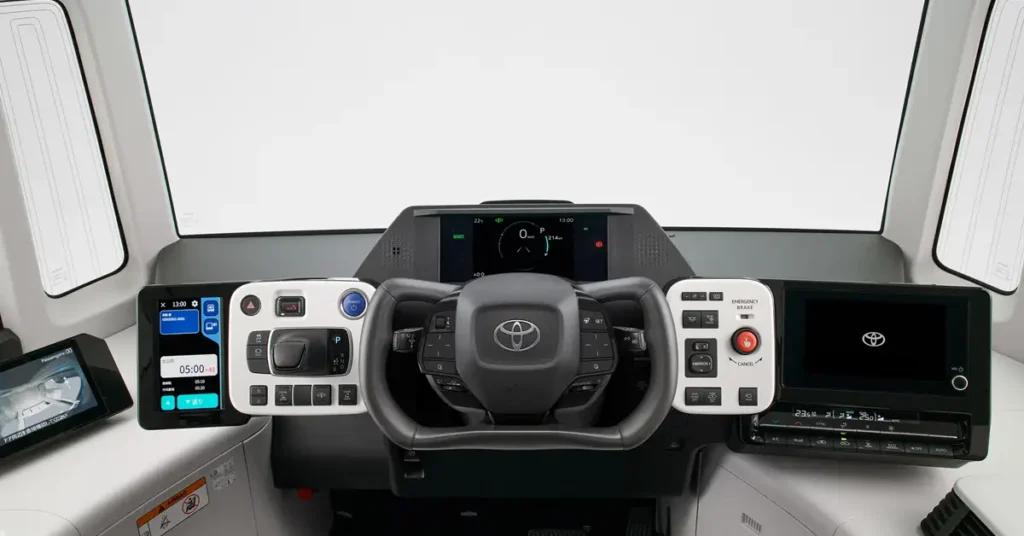 A high-angle, medium shot captures the interior of a Toyota e-Palette , focusing on the dashboard and steering wheel. The dashboard is predominantly white and gray, with a futuristic design featuring multiple screens and control panels.
