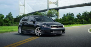 A medium shot captures a dark gray Volkswagen Golf parked on an asphalt road, with a large suspension bridge in the background.