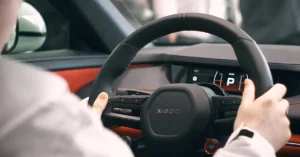 A close-up shot focuses on the steering wheel of a Xiaomi car, with a person's hands gently resting on it. The steering wheel is a sleek, matte black with the brand name "Xiaomi" subtly embossed in the center