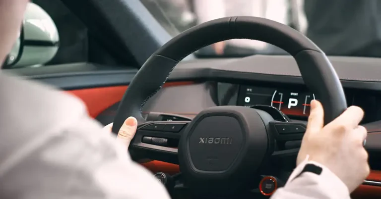 A close-up shot focuses on the steering wheel of a Xiaomi car, with a person's hands gently resting on it. The steering wheel is a sleek, matte black with the brand name "Xiaomi" subtly embossed in the center