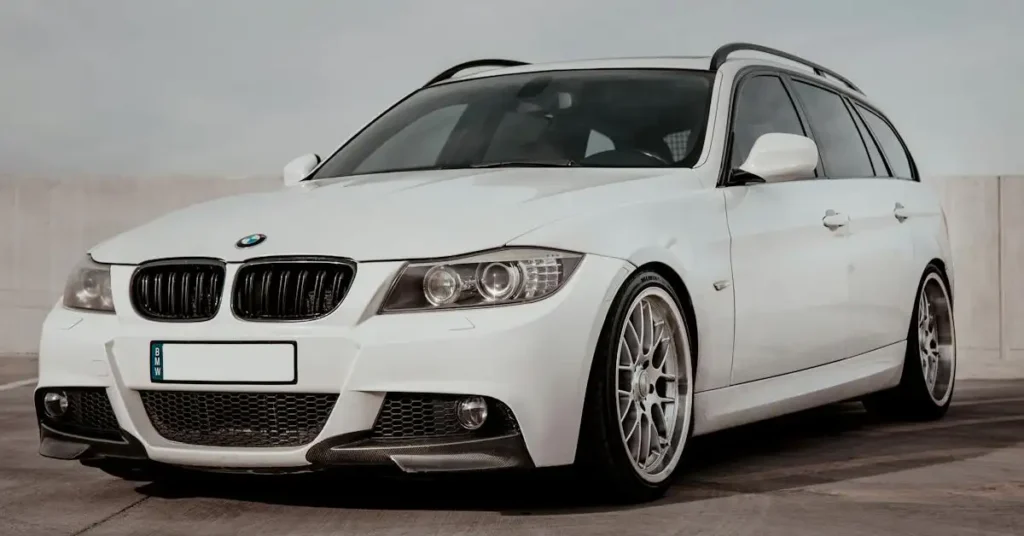 A white BMW 3 Series station wagon is parked on a concrete surface. The car has a black roof rack and silver wheels. The front of the car is visible, and the BMW logo is prominently displayed on the hood.