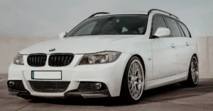 A white BMW 3 Series station wagon is parked on a concrete surface. The car has a black roof rack and silver wheels. The front of the car is visible, and the BMW logo is prominently displayed on the hood.