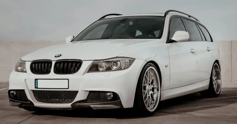 A white BMW 3 Series station wagon is parked on a concrete surface. The car has a black roof rack and silver wheels. The front of the car is visible, and the BMW logo is prominently displayed on the hood.