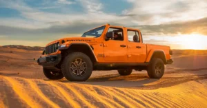 A bright orange 2025 Jeep Gladiator Mojave pickup truck is parked on a sand dune in a desert landscape. The truck is facing left, with its front wheels turned slightly.