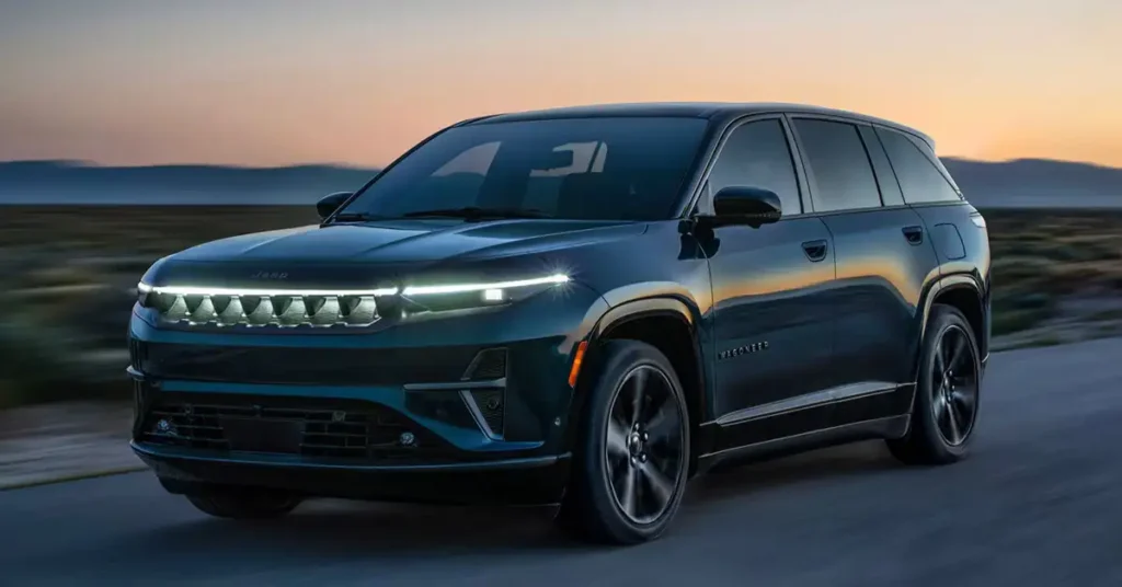 A sleek, dark blue 2025 Jeep Wagoneer S is captured in a dynamic, low-angle shot against a backdrop of a dusky sky and a blurred landscape. The vehicle is in motion, as suggested by the motion blur on the road beneath it. The front of the Jeep features a distinctive illuminated grille with the "Jeep" logo subtly integrated above it, flanked by modern, slim headlights.