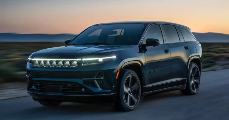 A sleek, dark blue 2025 Jeep Wagoneer S is captured in a dynamic, low-angle shot against a backdrop of a dusky sky and a blurred landscape. The vehicle is in motion, as suggested by the motion blur on the road beneath it. The front of the Jeep features a distinctive illuminated grille with the "Jeep" logo subtly integrated above it, flanked by modern, slim headlights.