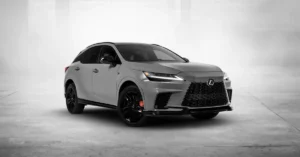 A 2025 Lexus RX  F Sport is shown in a studio setting. The SUV is a light gray color with black accents, including the grille, window trim, and wheels. The wheels are large and have a sporty design, with red brake calipers visible behind the spokes.