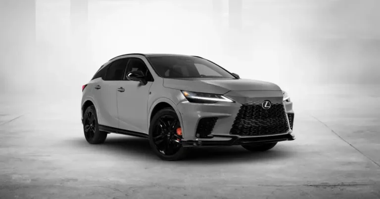 A 2025 Lexus RX  F Sport is shown in a studio setting. The SUV is a light gray color with black accents, including the grille, window trim, and wheels. The wheels are large and have a sporty design, with red brake calipers visible behind the spokes.