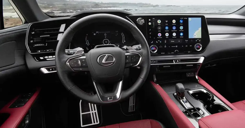 A wide-angle shot captures the interior of a modern 2025 Lexus RX car, featuring a luxurious red and black color scheme. The focal point is the steering wheel, adorned with the Lexus logo and various control buttons. Behind it, a digital dashboard displays driving information.