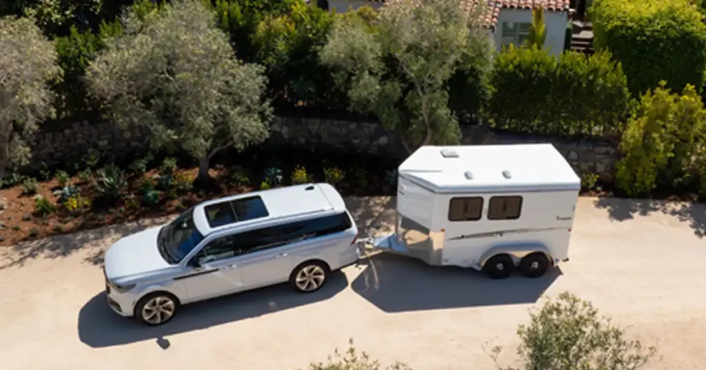An aerial shot shows a white 2025 Lincoln Navigator SUV towing a white horse trailer on a gravel driveway. The SUV is a 2020 Lincoln Navigator L, and it has a sunroof. The horse trailer is a two-horse trailer with two axles and two windows on each side. The driveway is surrounded by trees and bushes, and there is a stone wall in the background. The lighting is natural daylight, and the image has a slightly desaturated color palette