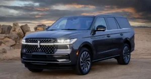 A dark gray 2025 Lincoln Navigator SUV is parked on a dirt road in a desert landscape. The SUV is facing the viewer at a slight angle, with its front left side more prominent. The vehicle has a large, intricate grille with a diamond pattern and a horizontal light bar that extends across the width of the front.