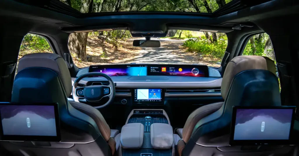 A wide-angle shot from inside a 2025 Lincoln Navigator SUV shows a panoramic view of the road ahead through the windshield and a large sunroof. The interior features two plush, tan leather seats in the foreground, with the backs of two dark blue leather seats visible behind them. Each of these rear seats has a built-in entertainment screen displaying the Lincoln logo.