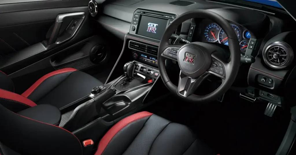A detailed shot of the interior of a 2025 Nissan GT-R , featuring a black and red color scheme. The seats are upholstered in black leather with red accents along the edges. The dashboard is predominantly black, with a central infotainment screen displaying the GT-R logo.