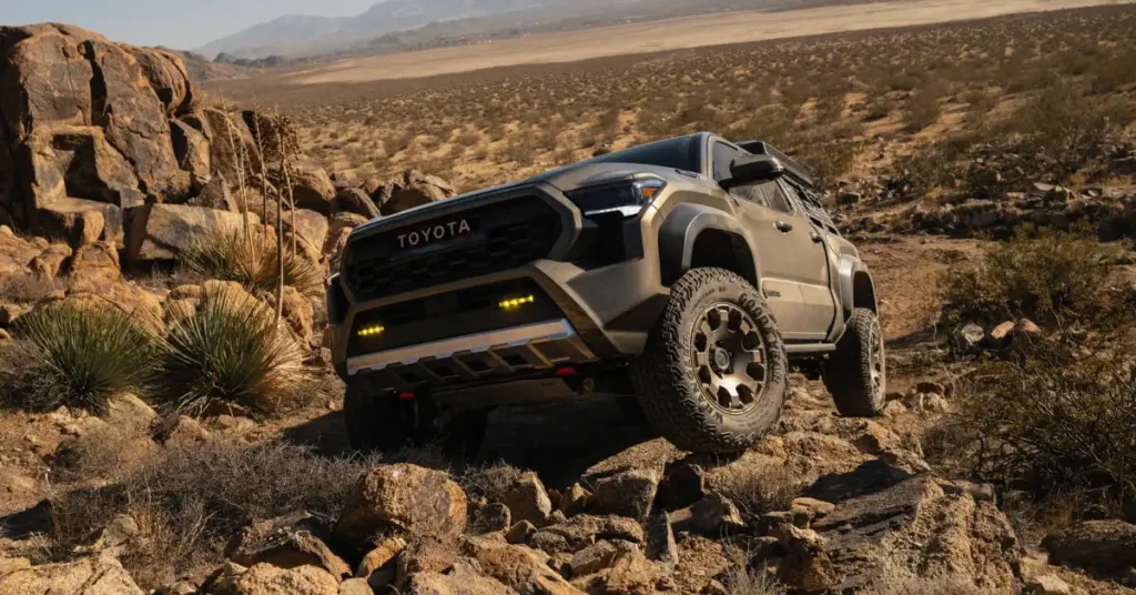 A tan Toyota 2025 Tacoma TRD Pro truck is shown from a low-angle, front-right perspective, navigating a rocky, desert terrain. The truck features a black grille with "TOYOTA" emblazoned across it, yellow fog lights, and aggressive off-road tires. The surrounding environment is arid, with sparse desert vegetation, large rocks, and distant hills under a clear sky.