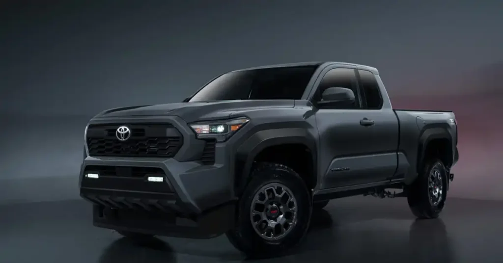A dark gray 2025 Toyota Tacoma TRD Pro pickup truck is shown in a studio setting. The truck is angled to the left, with its front end prominently displayed. It features a black honeycomb grille with the Toyota logo in the center, flanked by sharp, angular headlights. Below the grille, there are two horizontal LED light bars. The truck has rugged, dark gray fenders and off-road tires with distinctive TRD wheels.
