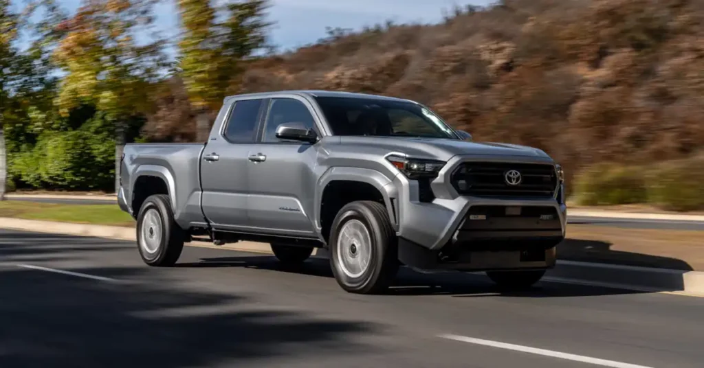 A silver Toyota 2025 Tacoma pickup truck is driving on a road. The truck is a four-door model with a crew cab and a short bed. It has a prominent grille with the Toyota logo in the center, and sharp, angular headlights. The truck's body is sleek and modern, with sculpted lines and flared wheel arches.