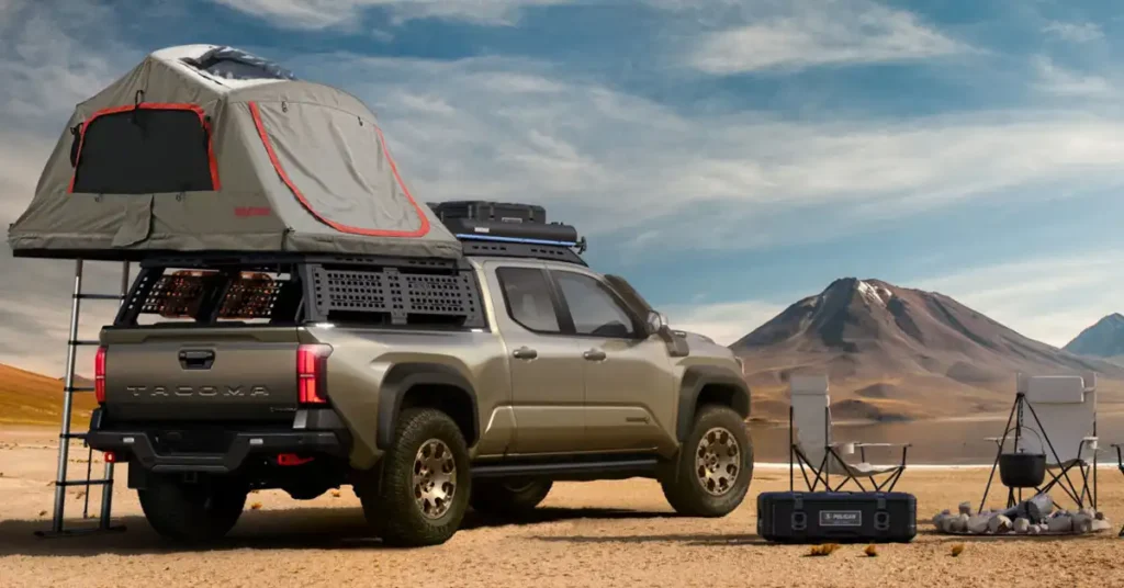A tan Toyota 2025 Tacoma pickup truck is parked on a sandy, arid landscape with mountains in the background. The truck is equipped with a rooftop tent, a cargo rack system in the bed, and off-road tires. In front of the truck, there are two camping chairs, a Pelican case, and a campfire with a pot hanging over it.