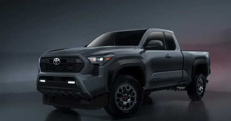 A dark gray 2025 Toyota Tacoma TRD Pro pickup truck is shown in a studio setting. The truck is angled to the left, with its front end prominently displayed. It features a black honeycomb grille with the Toyota logo in the center, flanked by sharp, angular headlights. Below the grille, there are two horizontal LED light bars. The truck has rugged, dark gray fenders and off-road tires with distinctive TRD wheels.