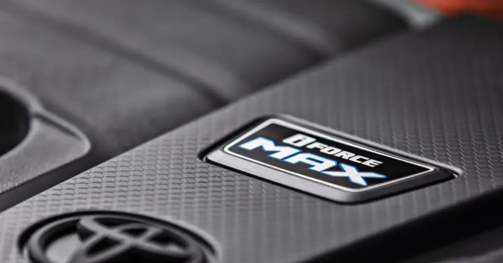 A close-up, low-angle shot shows the "i-FORCE MAX" emblem on a 2025 Toyota Tacoma TRD Pro textured, dark gray engine cover. The emblem is rectangular with a black border and a white outline. Inside, the words "i-FORCE" are in white, and "MAX" is in large, bold, blue letters with a white outline.