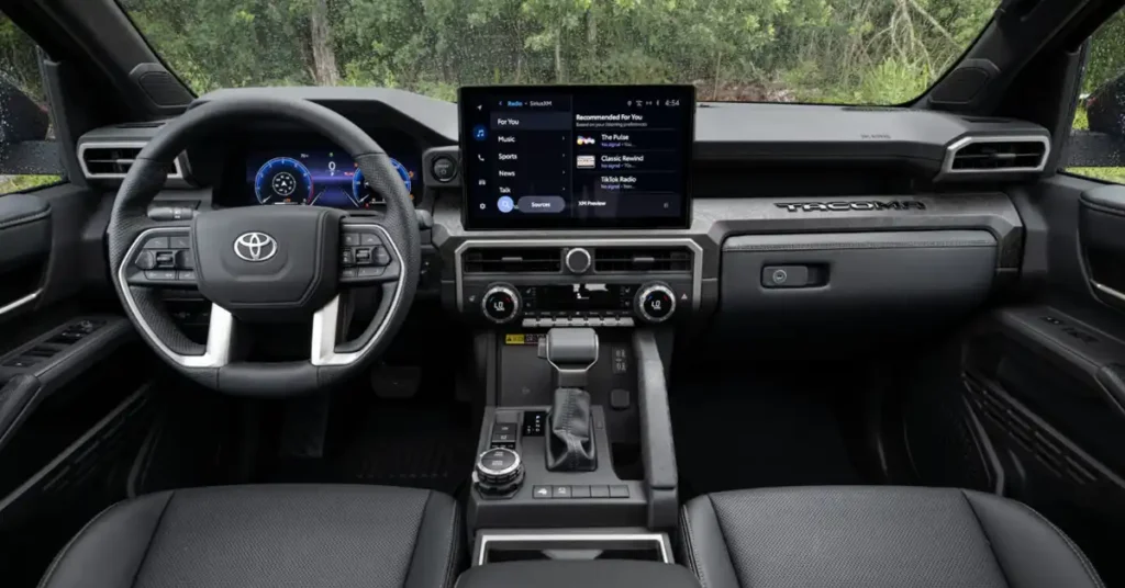 The image shows the interior of a Toyota Tacoma truck. The dashboard is dark gray with silver accents. The steering wheel is black leather with silver trim and a Toyota logo in the center. The instrument cluster is digital and displays blue and white graphics.