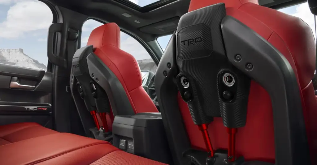 A close-up, eye-level shot shows the interior of a 2025 Toyota Tacoma truck, focusing on the front seats and the center console. The seats are upholstered in a vibrant red leather with black accents and carbon fiber detailing on the seatbacks.