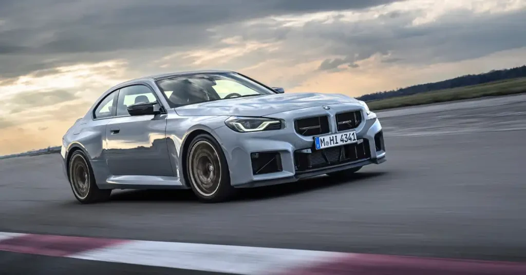 A silver BMW M2 coupe is captured in motion on a race track, with a dramatic, cloudy sky overhead. The car's sleek, aerodynamic design is emphasized by its low profile and aggressive front bumper.