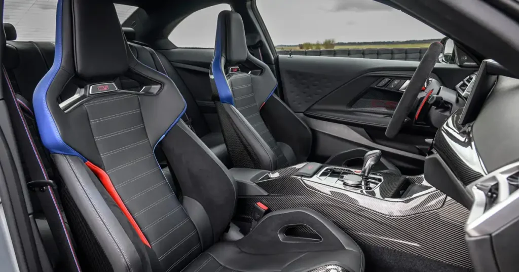 A close-up, eye-level shot captures the interior of a 2026 BMW M2 CS high-performance car, focusing on the front seats and center console. The seats are upholstered in black leather with contrasting white stitching and feature striking blue and red accents on the bolsters and headrests.