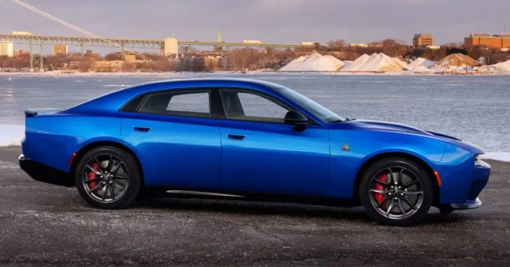 A side view of a blue 2025 Dodge Charger Daytona SRT electric muscle car parked on a road, with a body of water, a bridge, and buildings in the background. The car has a sleek, aerodynamic design with a long hood, a short rear deck, and a sloping roofline.