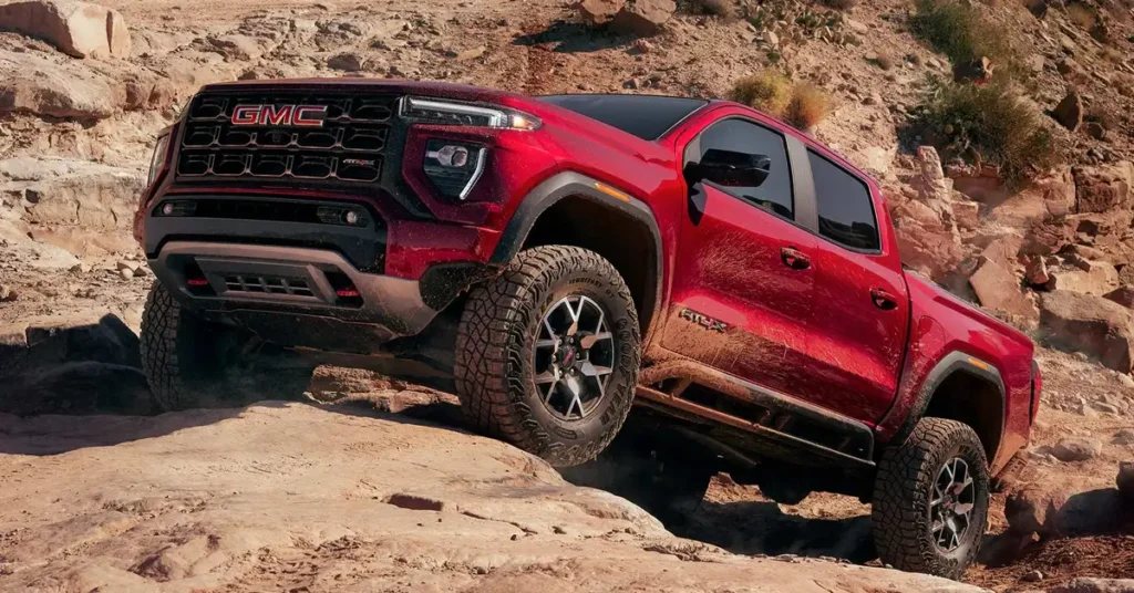 A red GMC Canyon AT4X pickup truck is shown climbing over rocks on a dirt road. The truck is angled slightly to the left, and the front of the truck is in focus. The truck has a black grille with the GMC logo in red, and the headlights are LED.