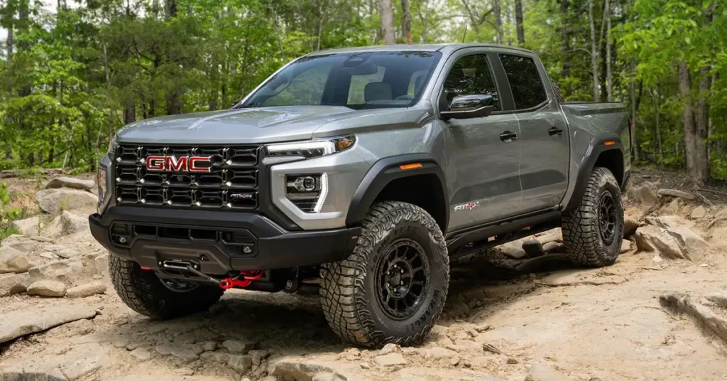 A gray 2026-GMC-Canyon pickup truck is shown on a rocky, dirt path in a forest. The truck is angled slightly to the left, showcasing its front and side. The front of the truck features a large, black grille with the GMC logo in red, flanked by modern LED headlights with a distinctive vertical daytime running light design.