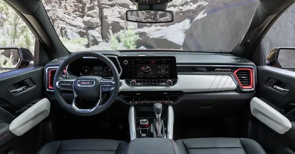 A photograph shows the interior of 2026 GMC Canyon pickup truck, focusing on the dashboard and front seating area. The dashboard features a large infotainment screen in the center, flanked by air vents and various controls. The steering wheel, adorned with the GMC logo, is positioned in front of the driver's seat.